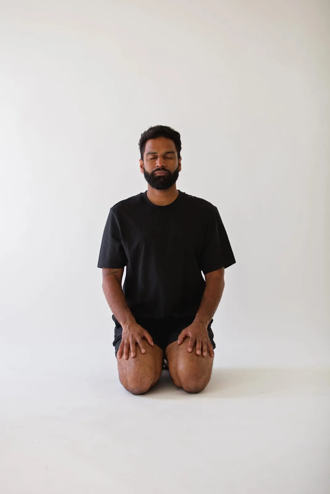 A man kneeling in quiet seated meditation, eyes closed, against a soft white backdrop